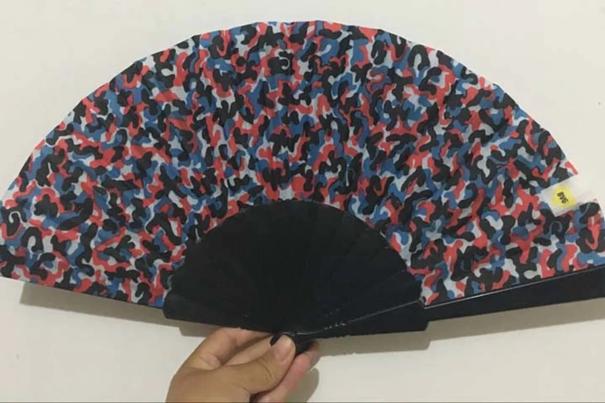 Multicolored printed folding fan with black ribs held open in one hand.