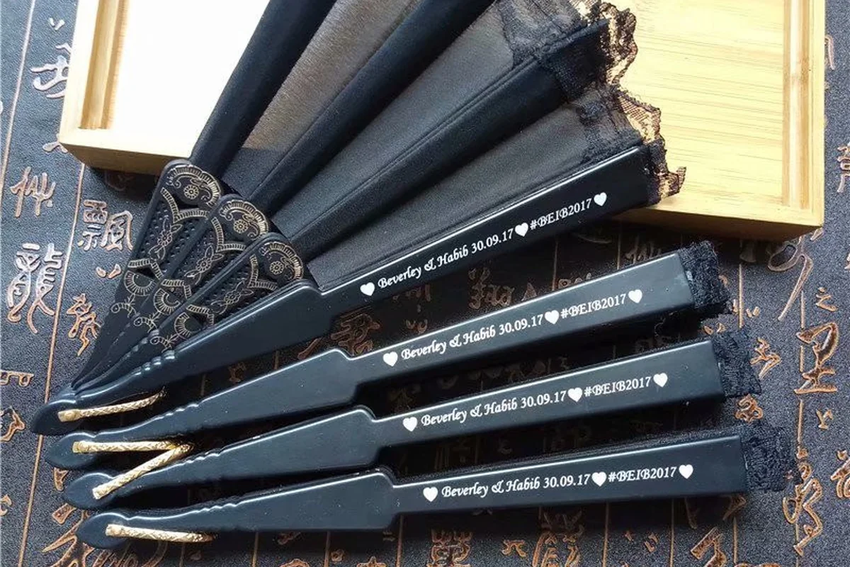 Folded black lace fans with lettering on their outer guards beside presentation boxes.